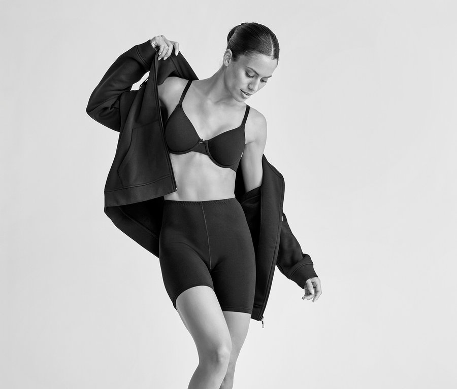 Eine Frau trägt einen schwarzen Bügel-BH und schwarze Shorts und zieht sich eine schwarze Kapuzenjacke an. Sie blickt nach unten, während sie die Jacke über ihre Schultern zieht.