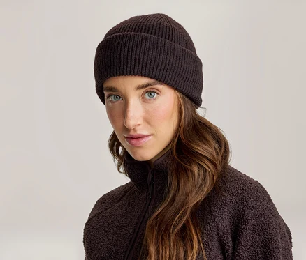 Frau mit langen braunen Haaren, die eine dunkelbraune gerippte Strickmütze und eine dunkelbraune Fleecejacke trägt und in die Kamera blickt.