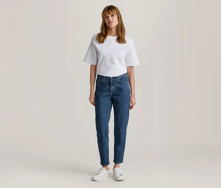 Frau steht, trägt ein weißes Kurzarm-T-Shirt, blaue Jeans und weiße Turnschuhe.