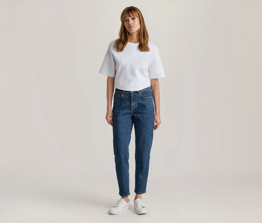 Frau steht, trägt ein weißes Kurzarm-T-Shirt, blaue Jeans und weiße Turnschuhe.