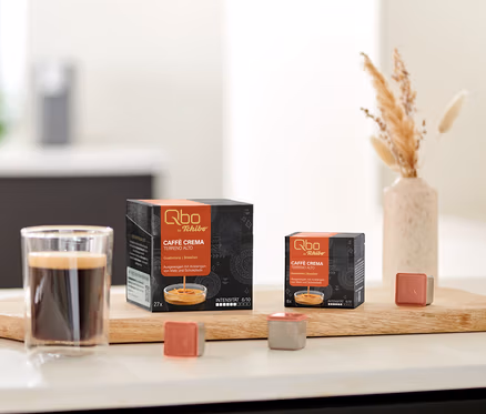 Ein Glas Kaffee steht auf einem Holzbrett neben zwei schwarzen Packungen Qbo Caffè TERRENO ALTO Kapseln, eine mit 27 und eine mit 8 Kapseln. Zwei einzelne Qbo Kapseln, eine rotbraune und eine graue, liegen vor den Packungen. Rechts steht eine Vase mit Trockengras, links eine Schale mit Keksen.