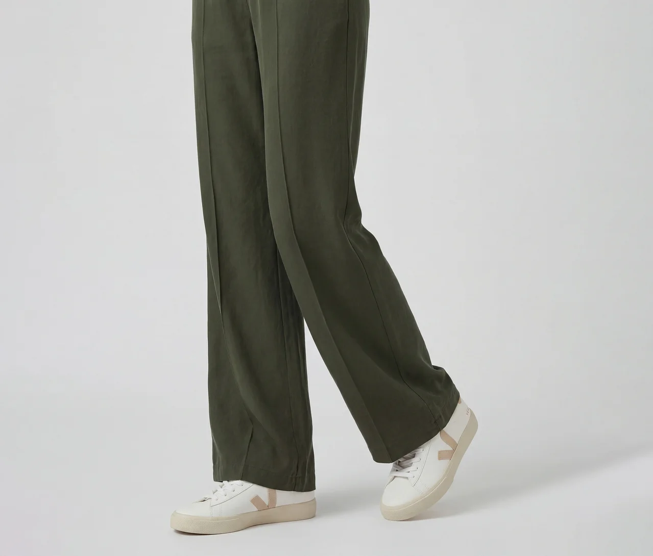Unterer Teil einer Person in dunkelgrüner weiter Hose und weißen Sneakern mit beigem V-förmigem Logo.