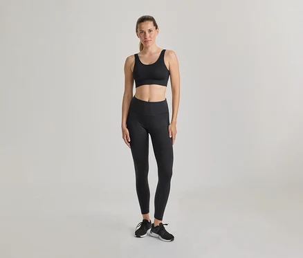Frau steht, trägt schwarzen Sport-BH und schwarze Leggings. Schwarze und weiße Turnschuhe, Haare zurückgebunden. Heller, einfarbiger Hintergrund.