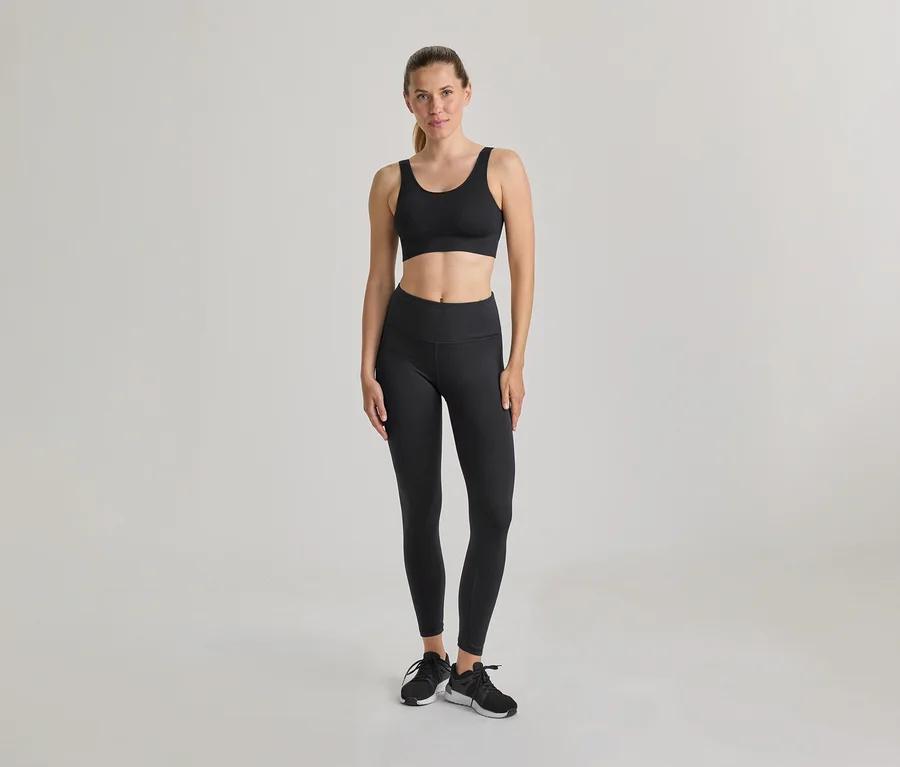 Frau steht, trägt schwarzen Sport-BH und schwarze Leggings. Schwarze und weiße Turnschuhe, Haare zurückgebunden. Heller, einfarbiger Hintergrund.