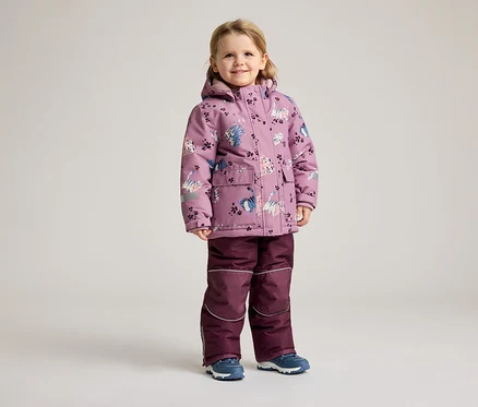 Ein kleines Mädchen steht in einem lila Wintermantel mit Katzenmuster und einer passenden Hose vor einem hellen Hintergrund.