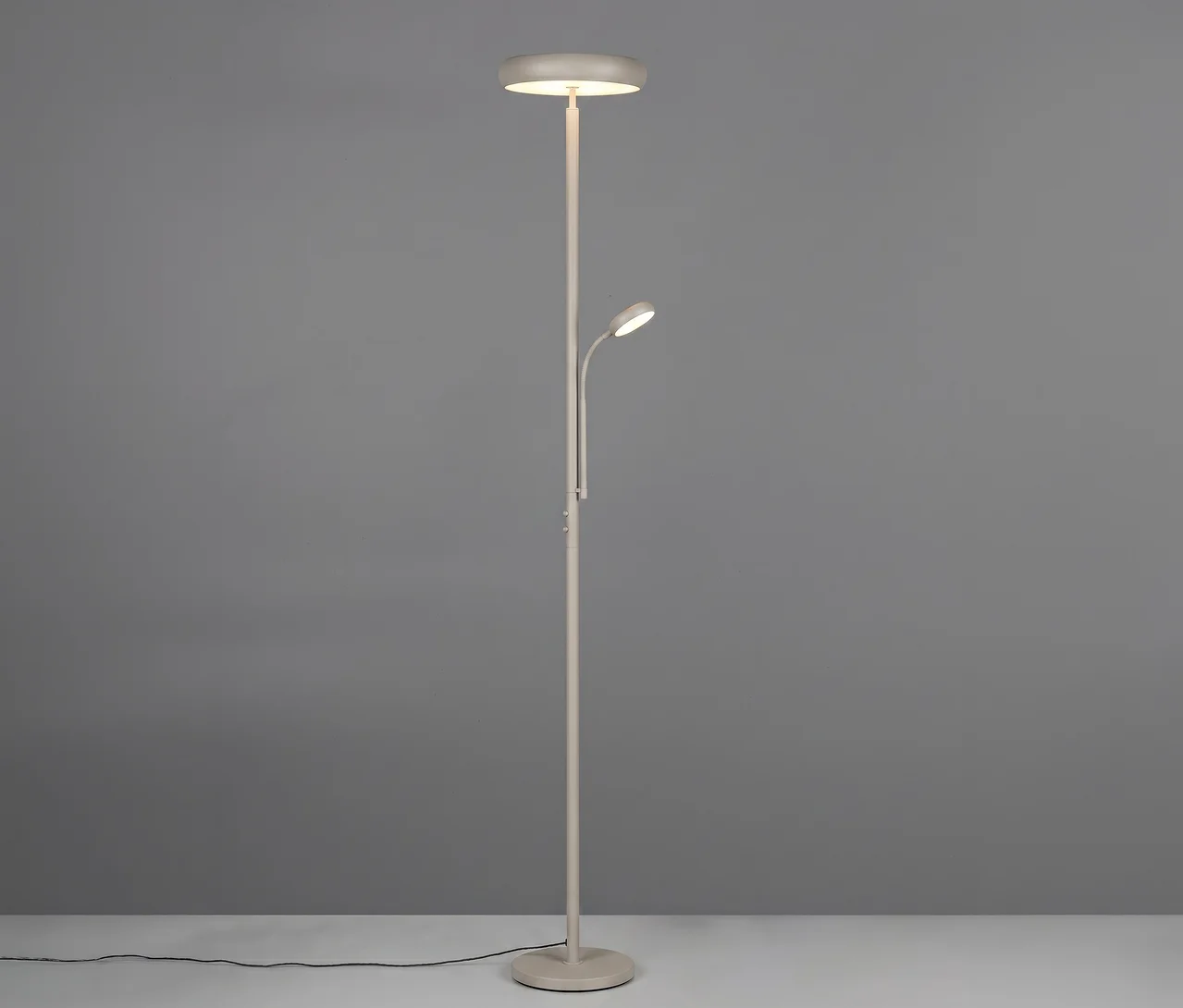 Eine hellgraue Stehleuchte mit zwei Lichtern. Das Hauptlicht ist ein Deckenfluter oben, und eine kleinere, verstellbare Leselampe ragt aus der Mitte des Ständers. Die Lampe steht auf einem hellen Boden vor einer grauen Wand, ein schwarzes Stromkabel ist auf dem Boden sichtbar.