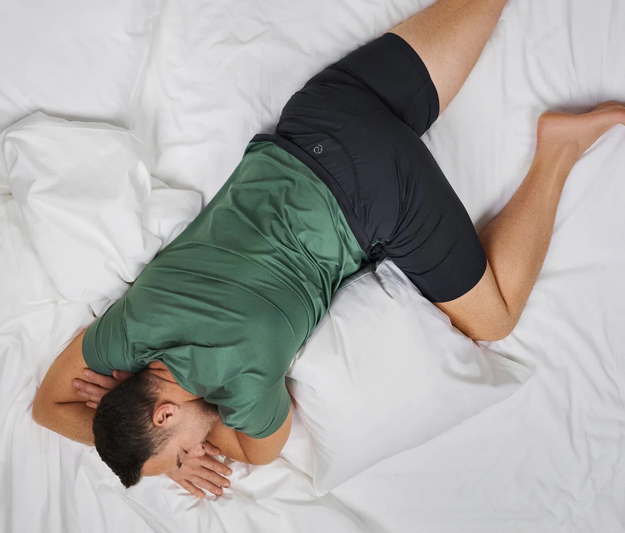 Mann schläft auf der Seite in einem CALIDA »Deepsleepwear Balancing« T-Shirt und CALIDA »Deepsleepwear Balancing« Shorts auf einem weißen Bett.
