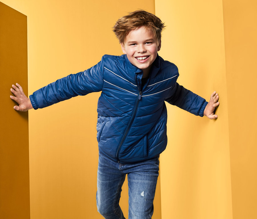 Ein Junge in einer blauen Steppjacke steht in der Ecke eines gelben Raumes.