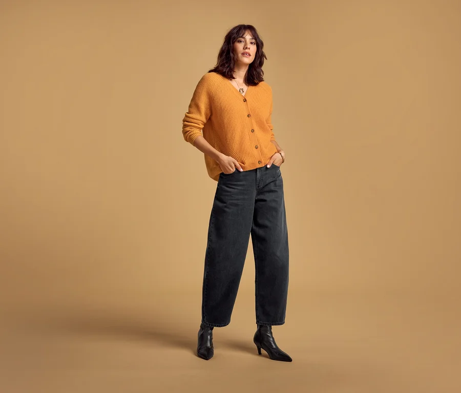 Eine Frau trägt einen orangefarbenen Merino-Cardigan und schwarze Barrel Jeans, die Hände in den Taschen.