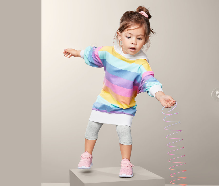 Mädchen steht auf einem grauen Würfel und hält ein Dehnspielzeug. Sie trägt ein Regenbogenkleid und 2 Leggings, 3/4-Länge.