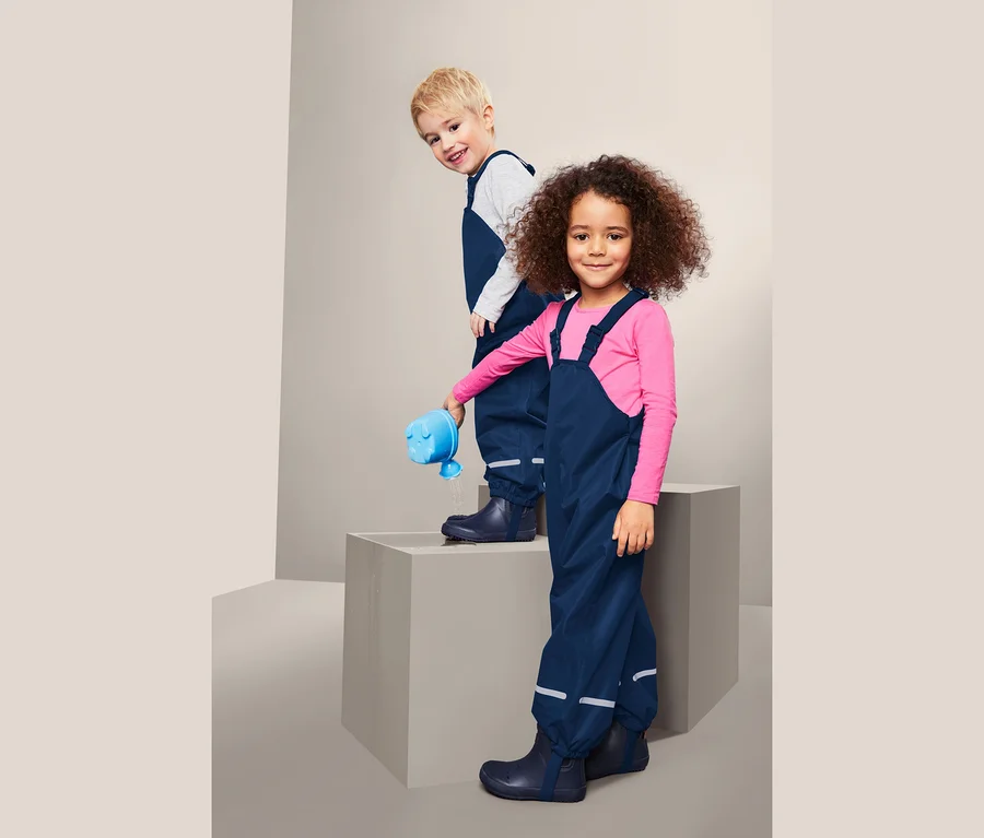Kinder tragen Regenhosen und spielen mit einer blauen Gießkanne.