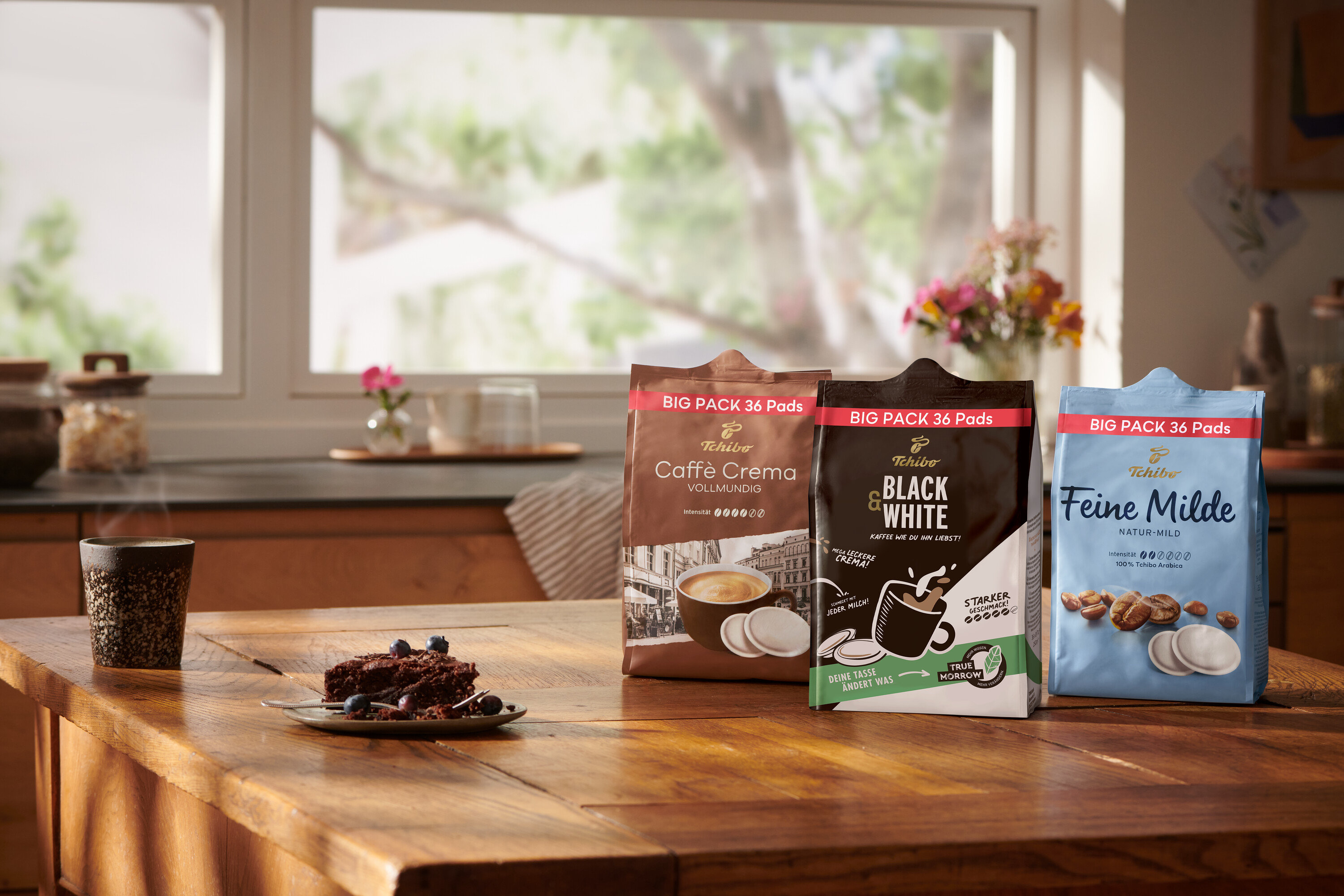 Auf einem Holztisch stehen eine Tasse, ein Teller mit Schokoladenkuchen und Packungen mit Tchibo Kaffeekapseln Caffè Crema, Black & White und Feine Milde.