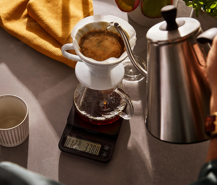 Person gießt Wasser aus einem elektrischen Schwanenhalskessel in eine Kaffeekanne aus Glas, die auf einer Kaffeewaage inkl. Timer steht.