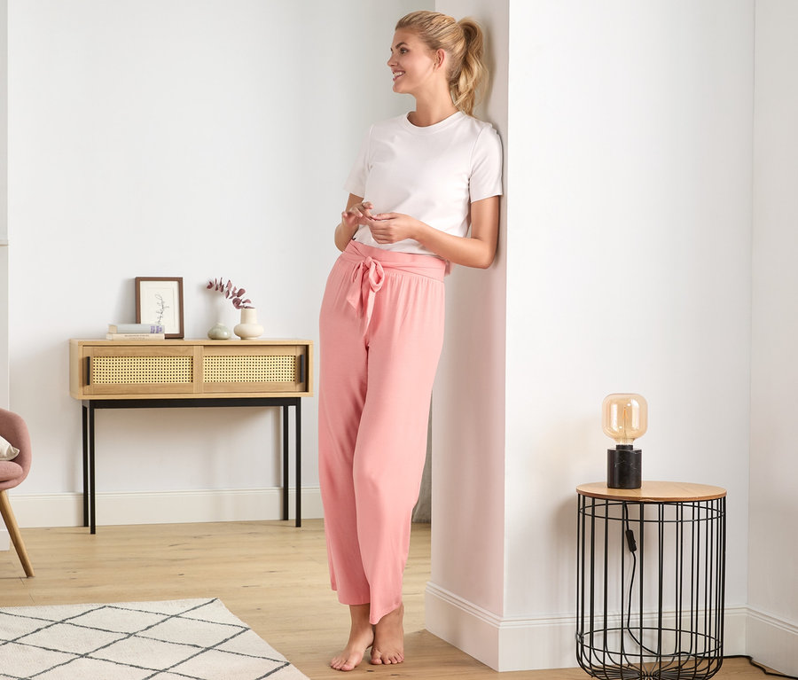 Blonde Frau lächelt und lehnt an einer Wand, sie trägt eine Relaxhose.