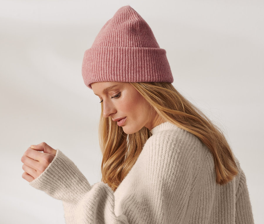 Blonde Frau mit einer roséfarbenen Strickmütze mit Wolle und cremefarbenem Pullover.
