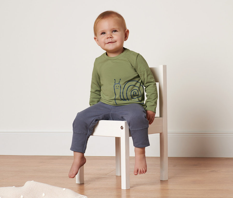 Ein Kind sitzt auf einem weißen Stuhl und trägt ein grünes Langarmshirt mit einem Schneckenaufdruck.