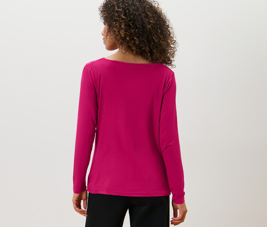 Rückansicht einer Frau mit braunen, lockigen Haaren, die ein Blusenshirt mit Wasserfallausschnitt und eine schwarze Hose trägt.