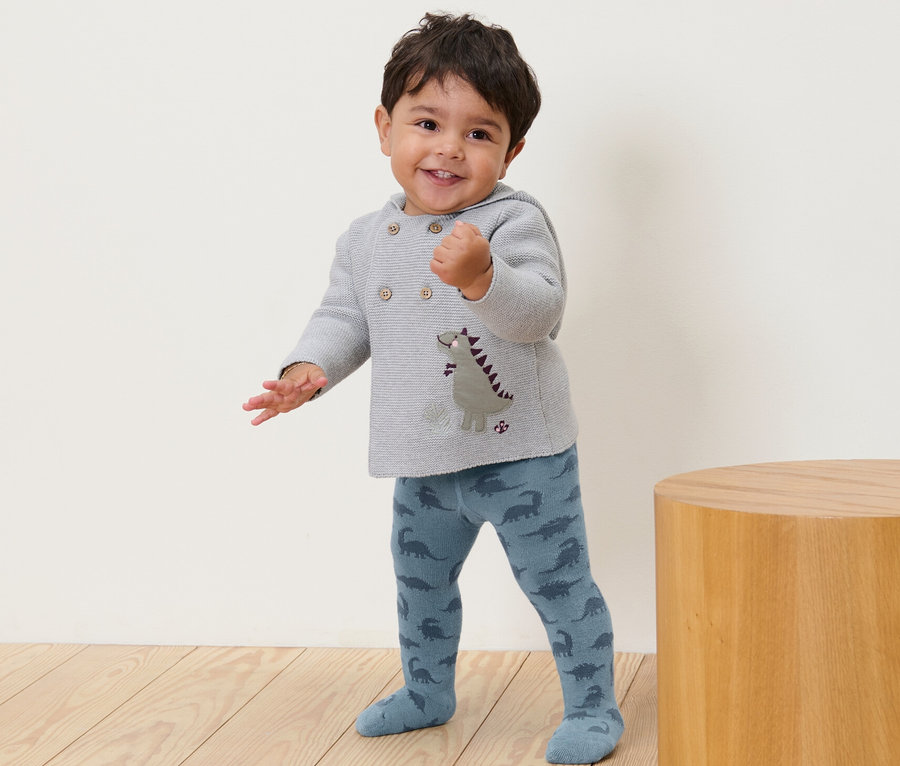 Ein lächelndes Kleinkind steht und trägt eine blaue Strumpfhose mit Dinosaurier-Muster.