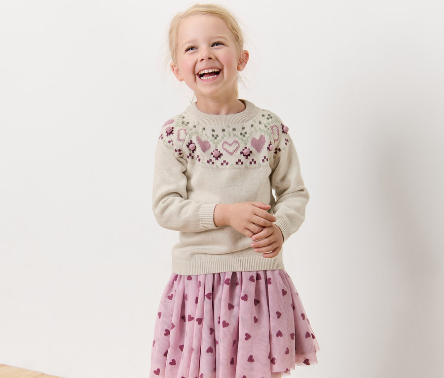 Lächelndes blondes Mädchen trägt einen Kinder-Strickpullover.
