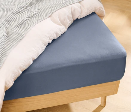 Blaues Jersey-Spannbettlaken auf einem Bett mit einer weißen Decke und einer grauen Strickdecke.