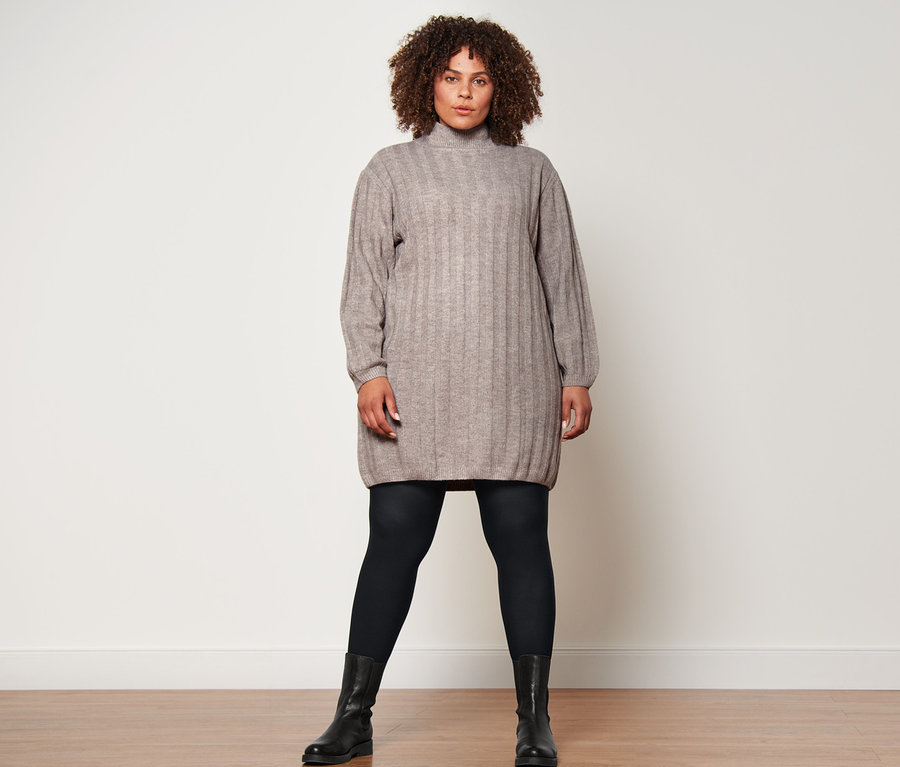 Eine Frau steht und trägt ein geripptes Strickkleid mit Stehkragen, eine schwarze, blickdichte Strumpfhose (2 Paar) und Lederboots.