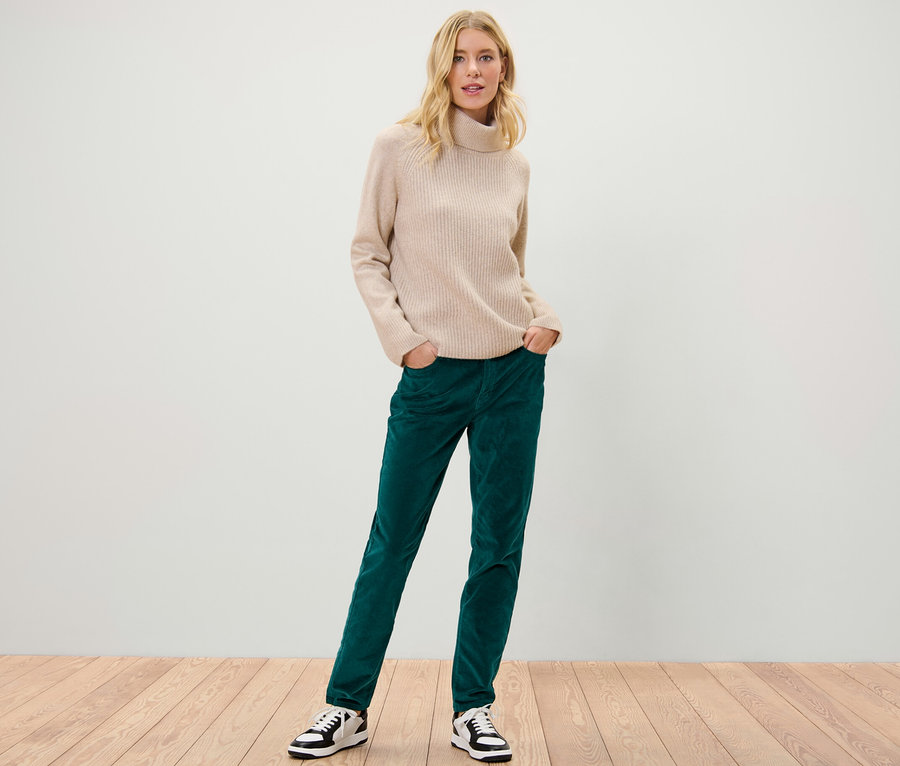 Blonde Frau posiert in petrolfarbener Samthose und beigem Rollkragenpullover.