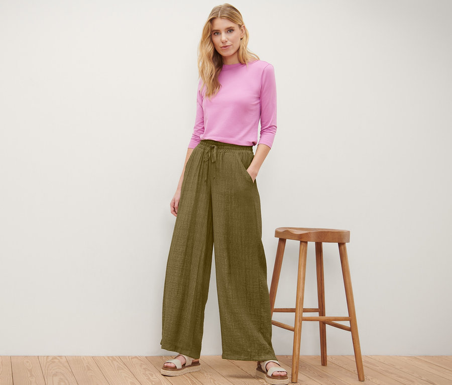 Frau posiert in olivfarbener Schlupfhose und pinkem Oberteil neben einem Holzhocker.