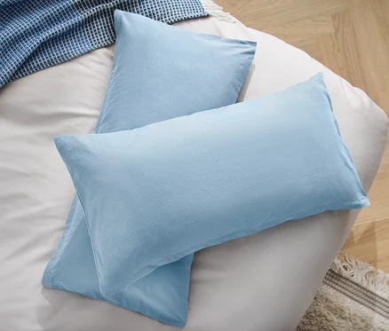 Zwei hellblaue Mikrofaser-Kopfkissenbezüge liegen auf einem Bett neben einer blauen Decke.