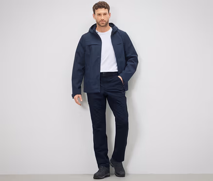 Mann posiert in Outdoorhose und Jacke vor einer Wand.