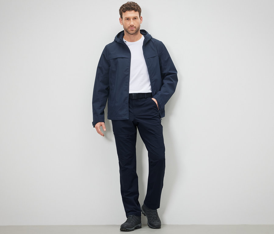 Mann posiert in Outdoorhose und Jacke vor einer Wand.