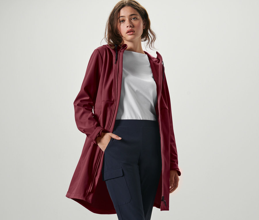 Frau posiert in weinrotem Softshellmantel, weißem T-Shirt und marineblauer Hose.