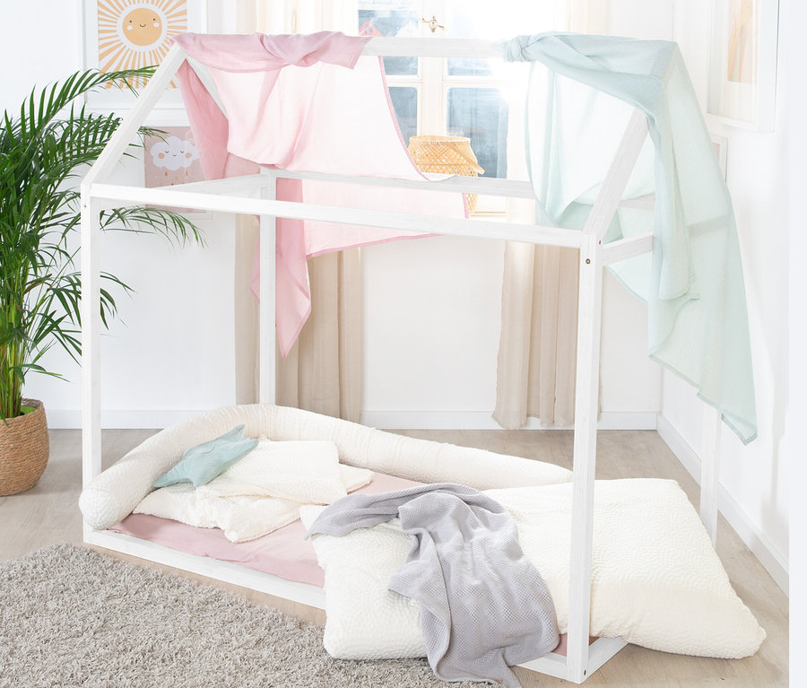 Ein weißes roba Hausbett, ca. 70 x 140 cm, mit rosa und blauen Stoffbahnen, die über den oberen Teil des Rahmens drapiert sind, steht auf einem Teppich.