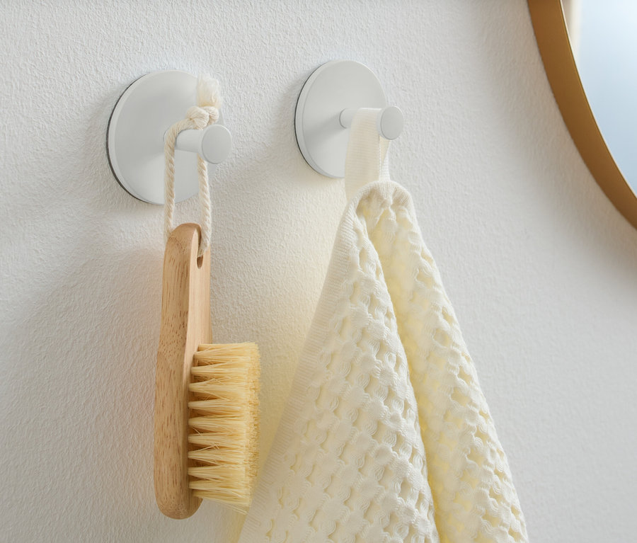 An einer weißen Wand hängen zwei Haken. An einem hängt eine Holzbürste und am anderen ein weißes Handtuch.