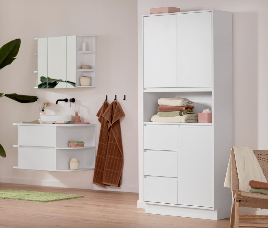 Badezimmer-Interieur mit weißem Bad-Hochschrank, Spiegelschrank und Waschbeckenunterschrank. Ein grüner Teppich und rostrote und cremefarbene Handtücher bringen Farbe ins Badezimmer.