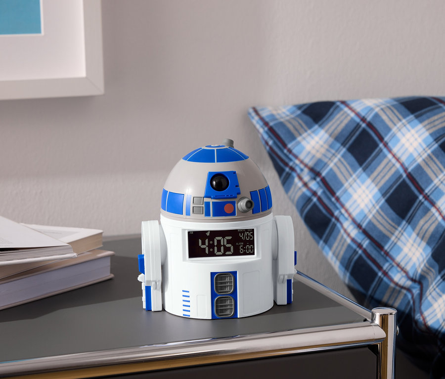 R2-D2 Wecker steht auf einem Nachttisch neben Büchern und einem blau-weiß karierten Kissen.