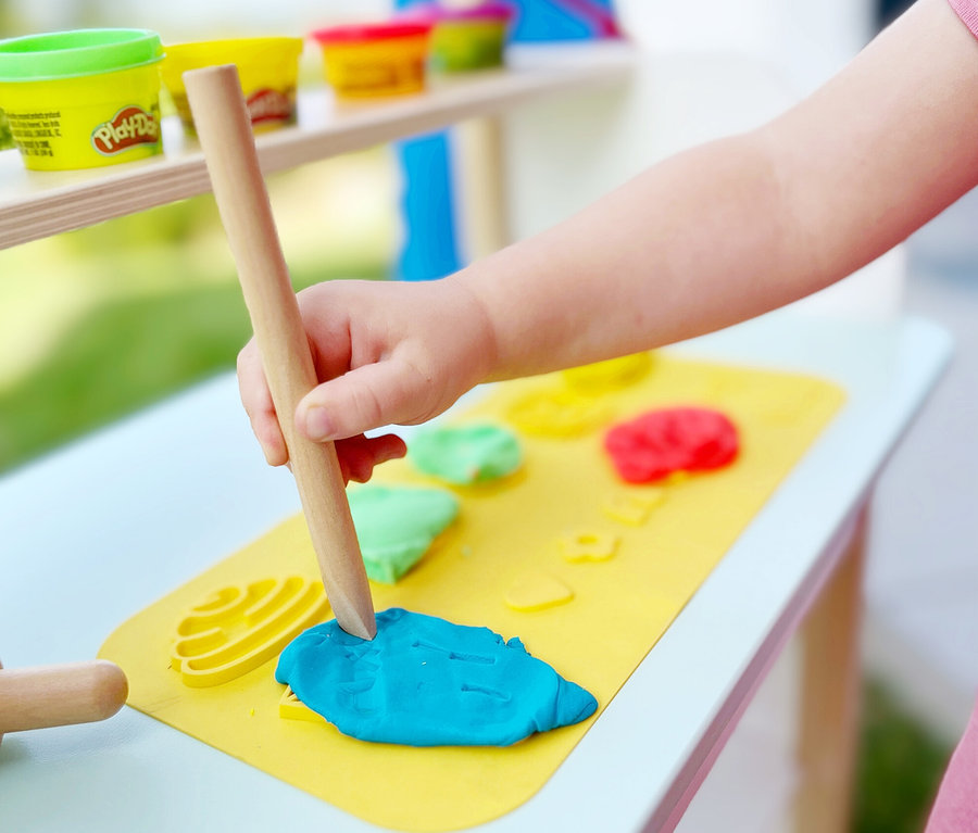 Kind rollt blaue Knete von roba x Play-Doh Knet-Spielset 1 mit einer Holzrolle auf einer gelben Matte aus. Auf der Matte liegen auch eine gelbe Form und weitere Knete.