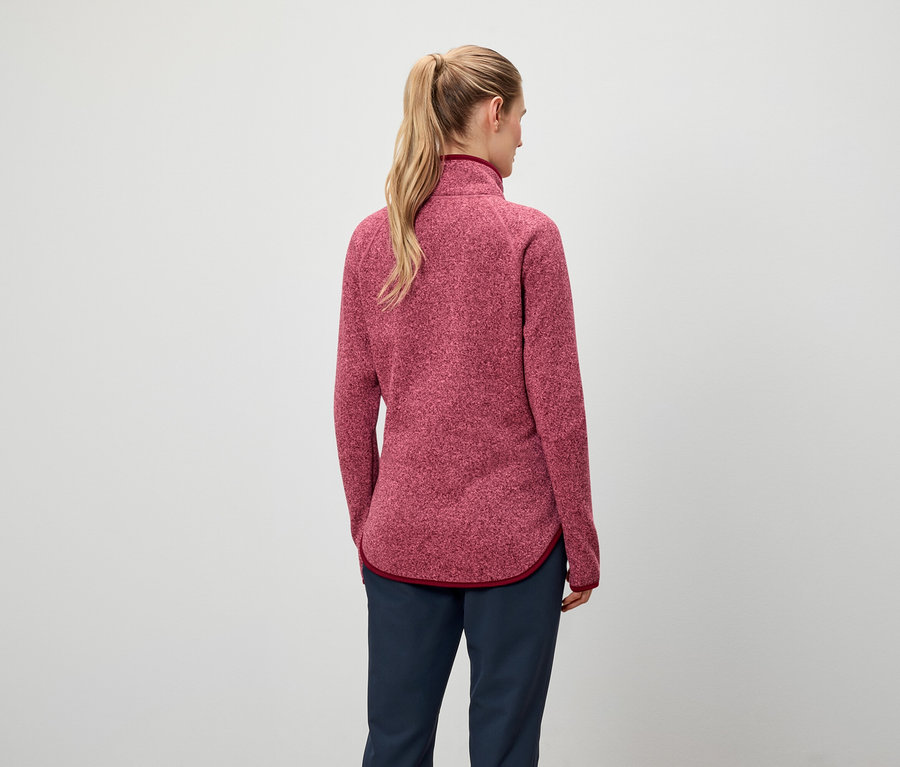 Von hinten ist eine Frau mit blonden, zu einem Pferdeschwanz zusammengebundenen Haaren zu sehen, die eine rote Sweatshirtjacke und dunkelblaue Hosen trägt.