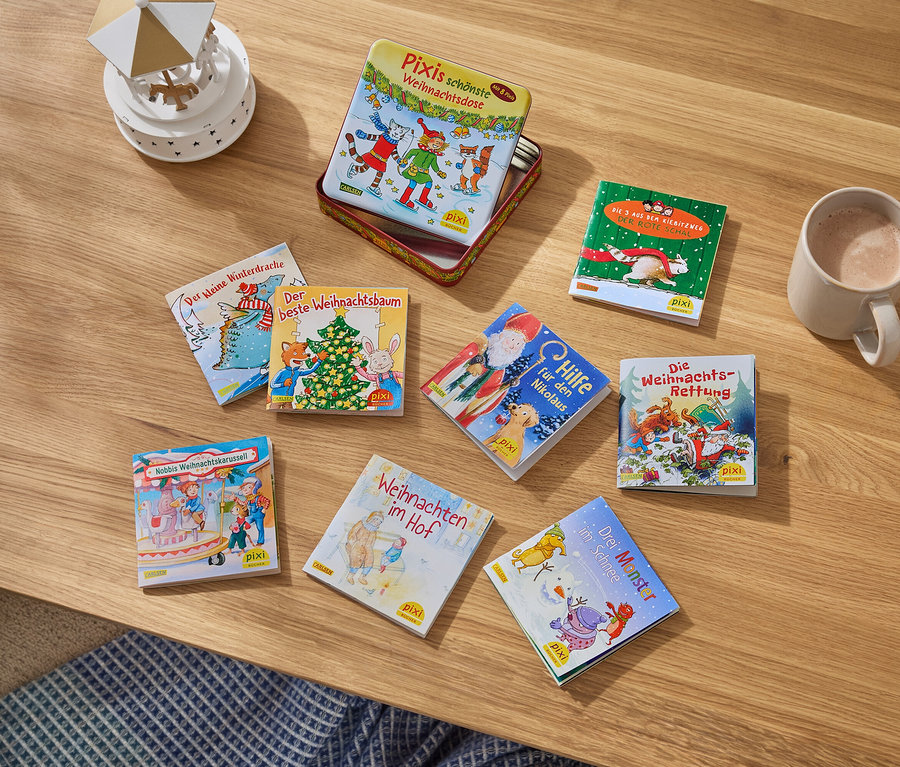 Auf einem Holztisch liegen Kinderbücher, eine Tasse mit einem Getränk und eine kleine Dekoration.