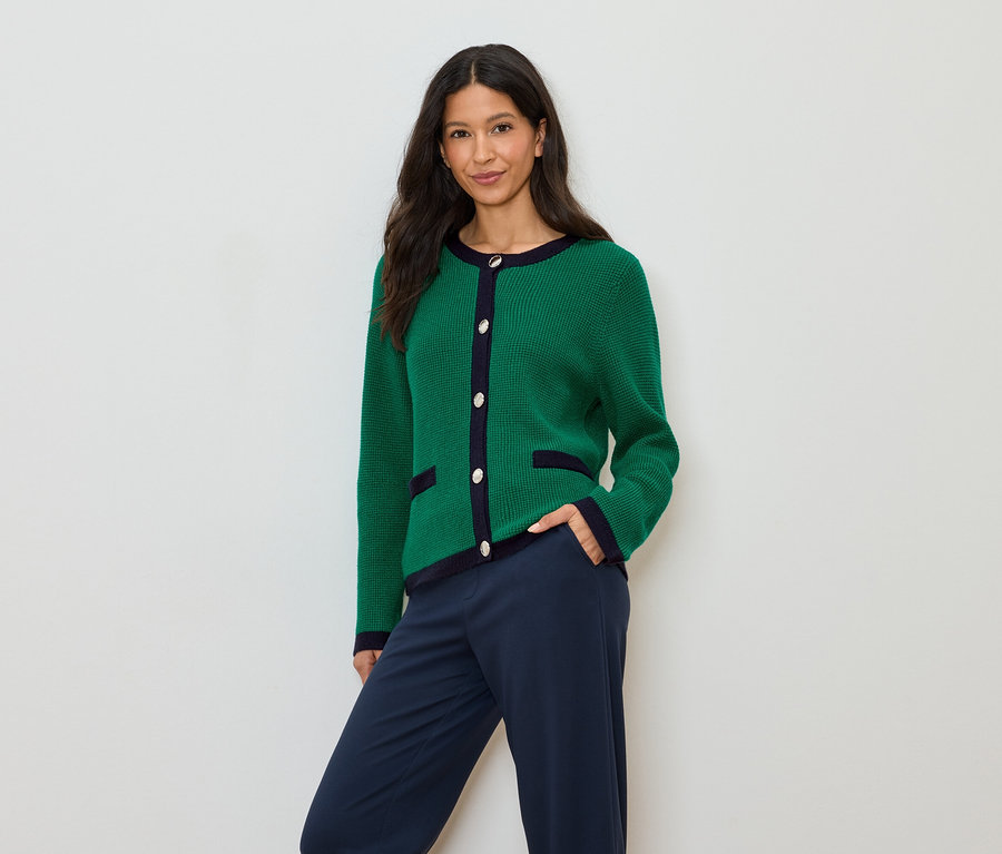 Eine Frau mit dunklen Haaren trägt eine grüne Strickjacke mit marineblauen Akzenten und marineblaue Hosen.