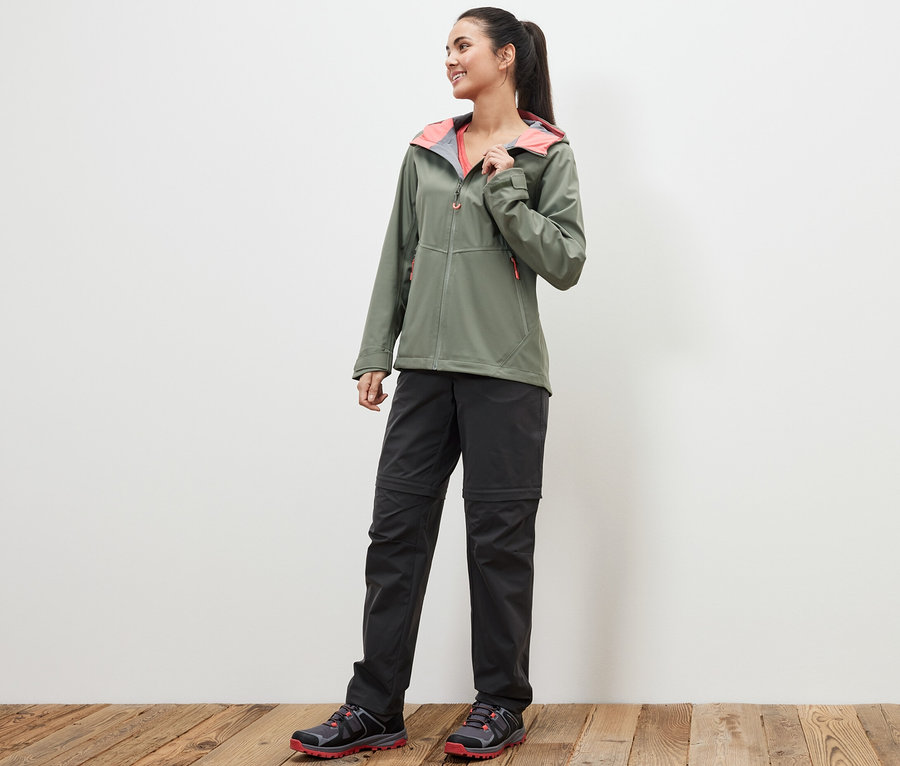 Eine Frau steht auf einem Holzboden und trägt eine grüne Jacke und eine schwarze 2-in-1-Funktionshose.