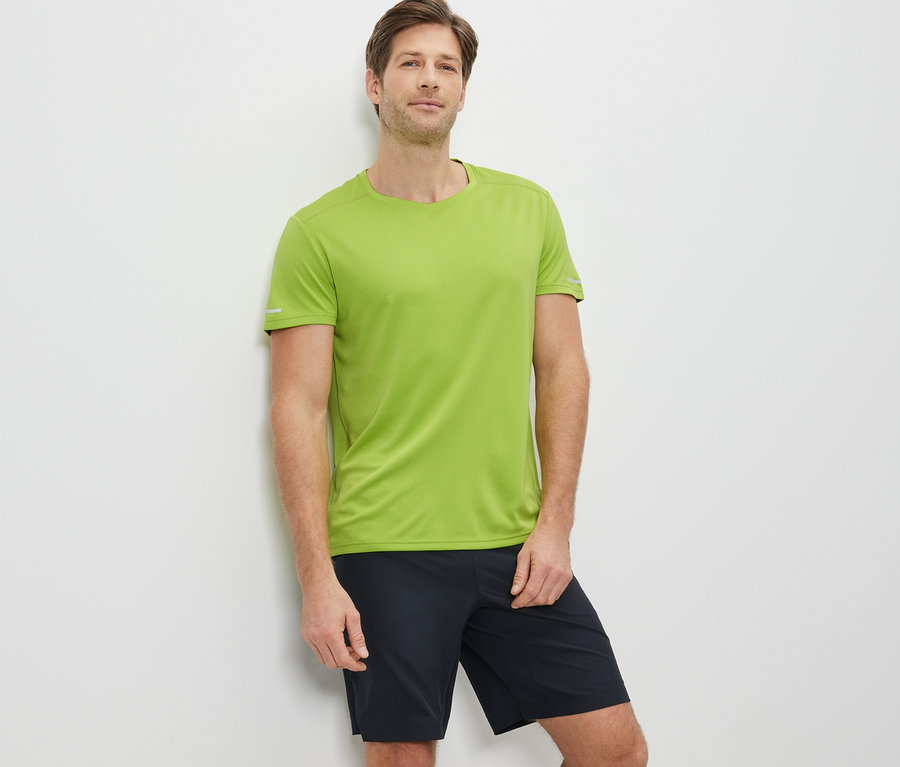 Mann lehnt an der Wand und trägt ein limefarbenes Funktionsshirt und schwarze Shorts.