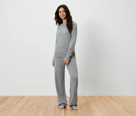 Eine Frau steht auf einem Holzboden und trägt einen grauen Strickpullover und eine Hose.