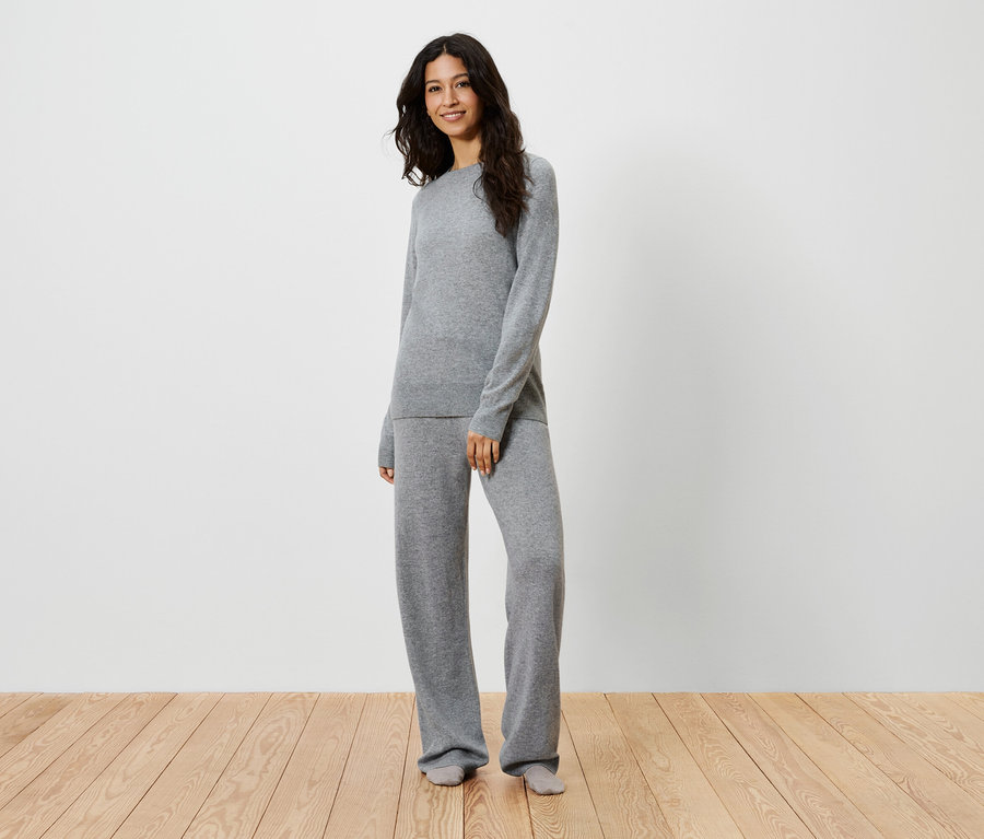 Eine Frau steht auf einem Holzboden und trägt einen grauen Strickpullover und eine Hose.