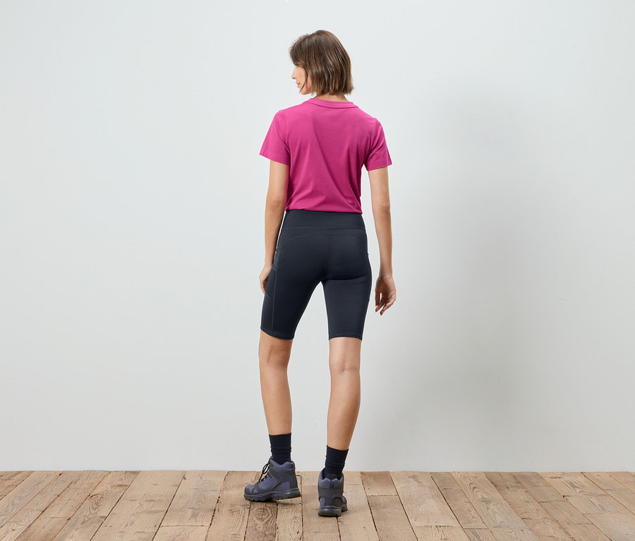 Rückansicht einer Frau mit kurzem braunen Haar, die ein kurzärmeliges, pinkfarbenes T-Shirt, kurze, marineblaue Leggings, marineblaue Socken und graue Wanderschuhe trägt.