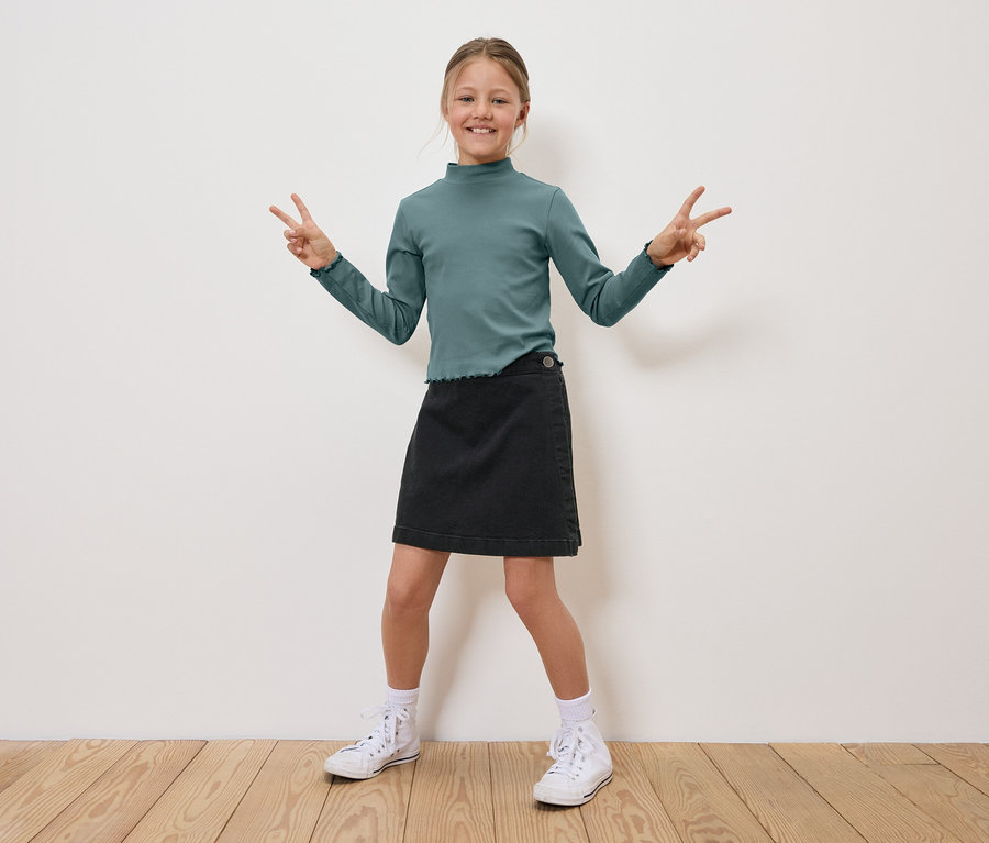 Ein Mädchen in einem grünen Oberteil und einem schwarzen Rock steht und zeigt mit den Fingern ein 'V'.