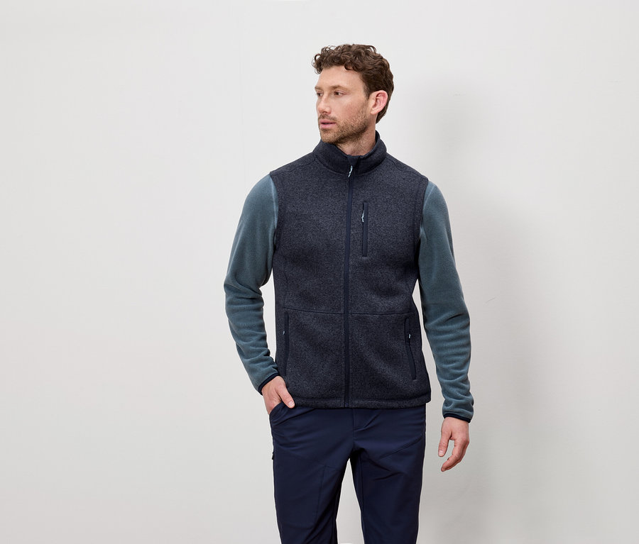 Mann mit kurzen lockigen Haaren trägt eine marineblaue Fleeceweste und ein blaugrünes langärmeliges Hemd und posiert vor einem hellen Hintergrund.