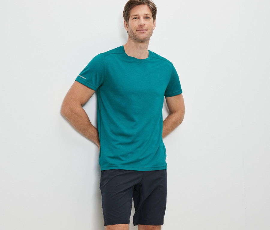 Mann lehnt an einer weißen Wand, trägt ein türkises Funktionsshirt und schwarze Shorts.
