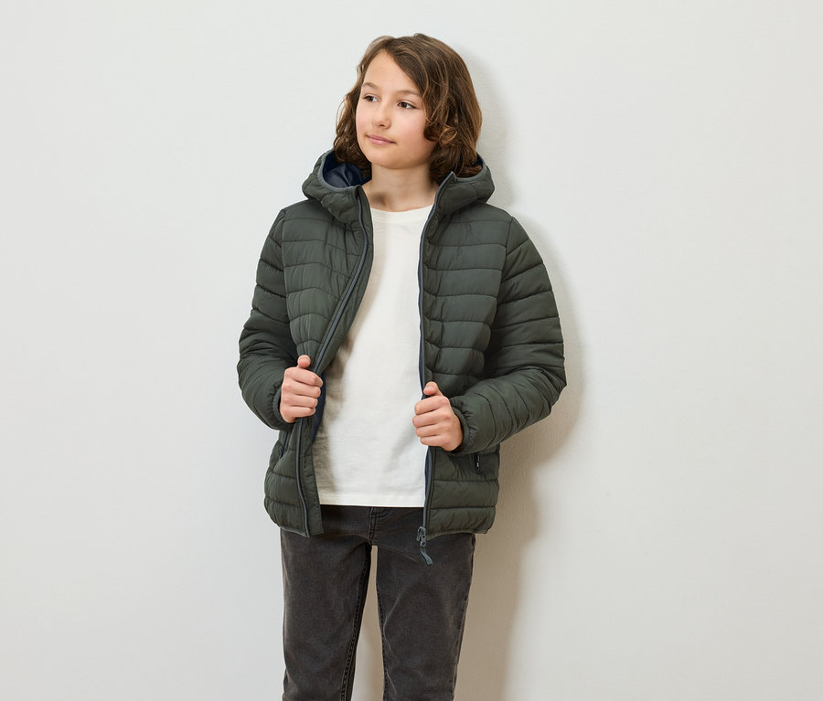 Eine jugendliche Person trägt eine olivgrüne Steppjacke mit Kapuze und schliesst sie. Die Person trägt Jeans und ein T-Shirt.