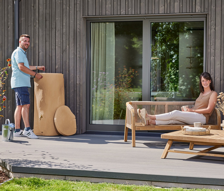 Ein Mann steht neben einem hellbraunen Regenwassertank auf einer Terrasse. Eine Frau entspannt sich auf einer Bank mit einem Getränk.