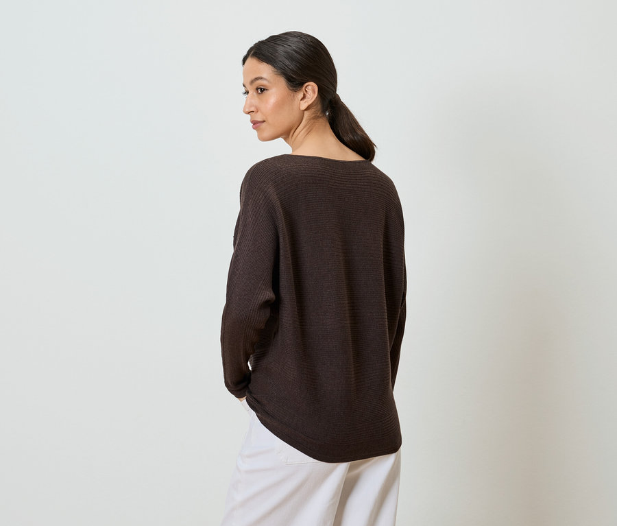 Eine Frau mit Zopf trägt einen braunen Pullover und weiße Hosen, die Hände in den Hosentaschen.